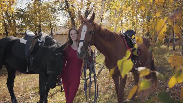 Smiling Caucasian Female Equestrian Holding Bridles of Two Horses and Looking Away. Young Girl in