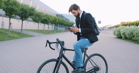 Bearded Man Sitting on Bike During Stop on the Pedestrian Path on the City Building Background