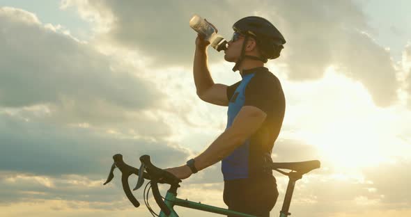 The Cyclist is Standing on the Edge of the Road and Drinking Water From a Bottle Sunset in the