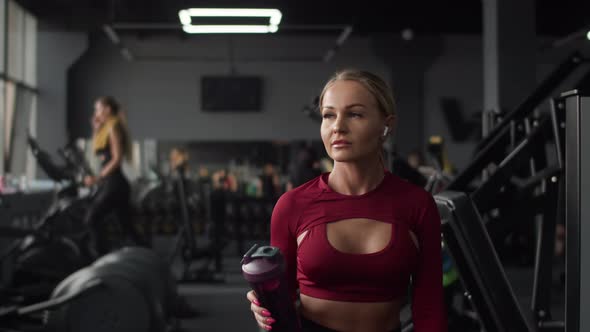 Beautiful Athletic Woman with Headphones with Bottle in Hand Walks Through Gym to Treadmill for