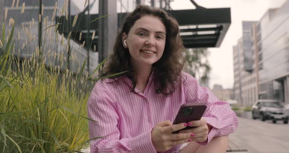 Portrait of joyful woman wearing pink shirt using a mobile phone, sit at the bus stop.