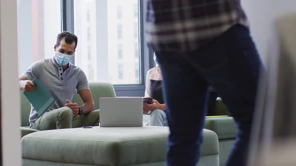 Diverse male and female business colleagues wearing face masks sitting on sofa discussing