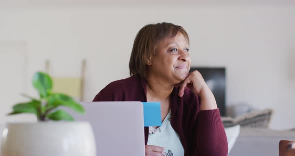 Happy african american senior woman at dining table using laptop, looking away and smiling