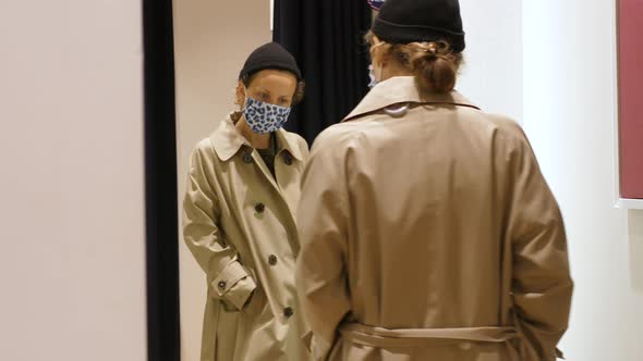 Young Woman Wearing Face Mask Trying Trench Coat in Fitting Room During Covid19