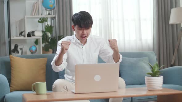 Happy Asian Man Celebrating While Using Laptop Computer In The Living Room