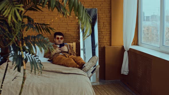 Relaxed Man in Bright Glasses Lying on Bed with Mobile Phone in Room