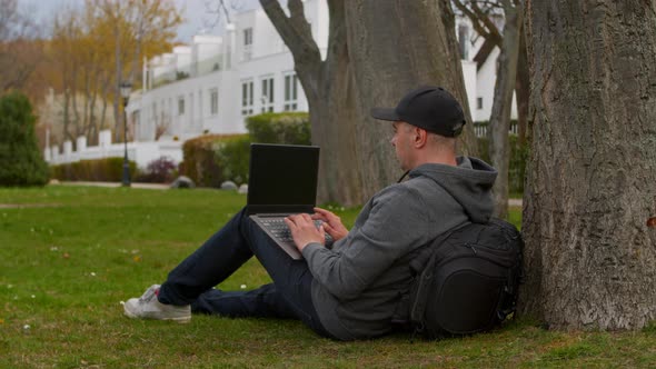 Young Man is Sitting in a Park in a Tourist Place Working on a Laptop