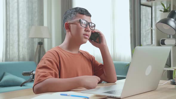 Asian Boy Sitting In A Wheelchair While Using Laptop Computer And Talking On Mobile Phone At Home