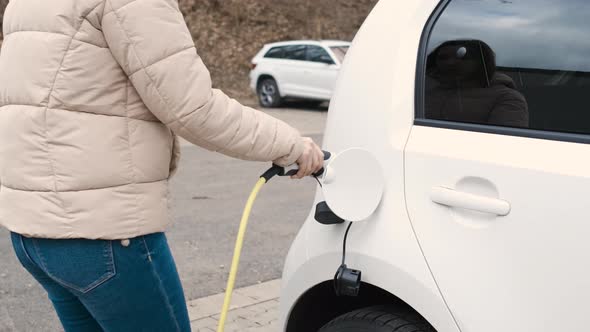 Woman Coming to the Electric Vehicle and Plugging in Power Supply Cable to Charge in the Street at