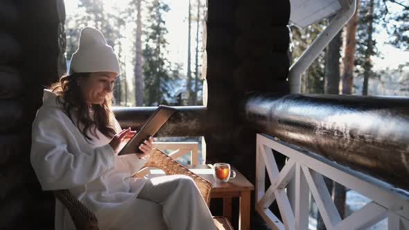 An Attractive Girl in Warm Clothes Sits on the Balcony and Works on the Tablet