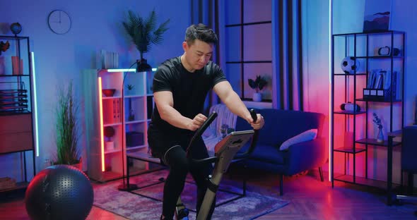 Asian Guy in Sportswear Exercising on Stationary Bike During Evening Home workout