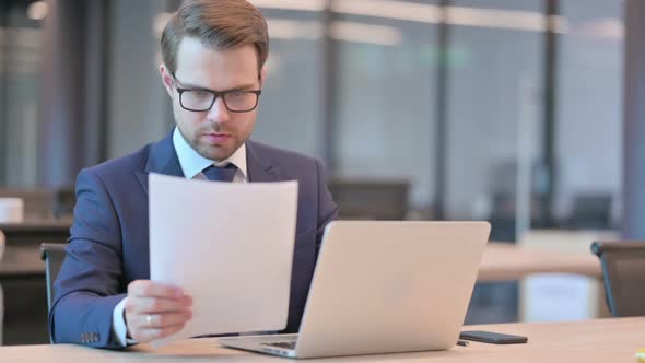 Businessman with Laptop Reading Documents in Office