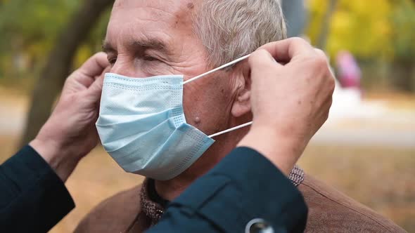 Senior Caucasian Woman Helps Puts a Medical Mask on the Face of an Senior Man