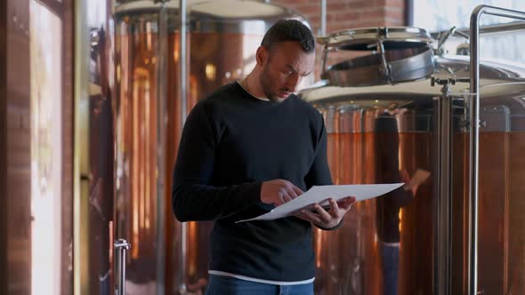 Serious Focused Man Standing with Paperwork in Brewery Looking at Large Beer Tanks