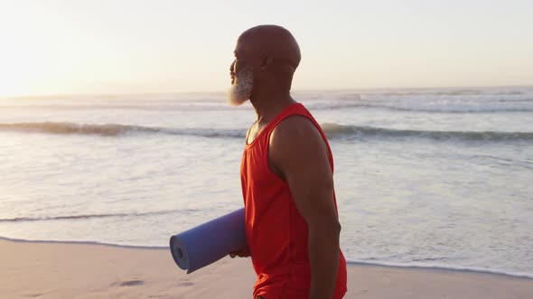 Senior african american man walking with yoga mat at the beach