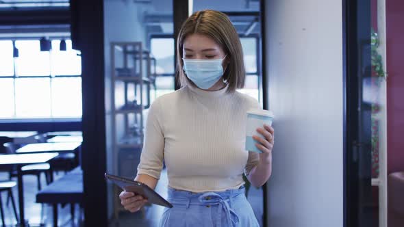 Asian businesswoman wearing mask walking using digital tablet in modern office