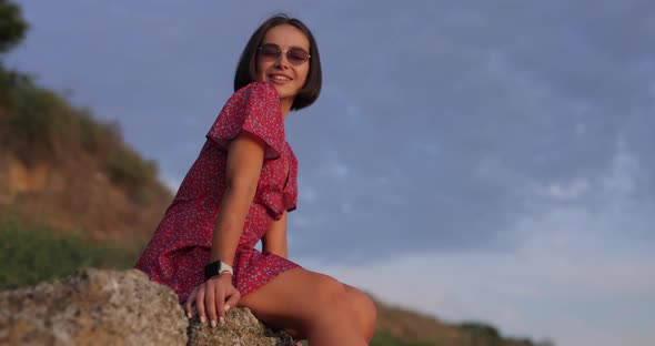 Happy Woman Sitting on a Stone Against a Sky Background in Sunglasses