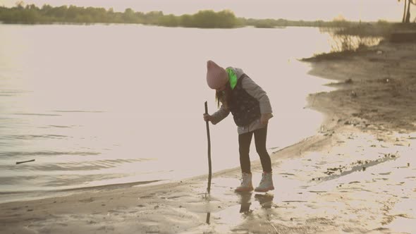 A Little Girl Plays with a Stick Alone at Sunset on the Shore of a Lake in Slow Motion