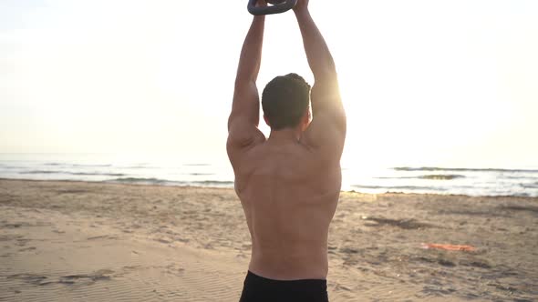 Shirtless man training with kettlebell on the beach at sunrise in slow motion