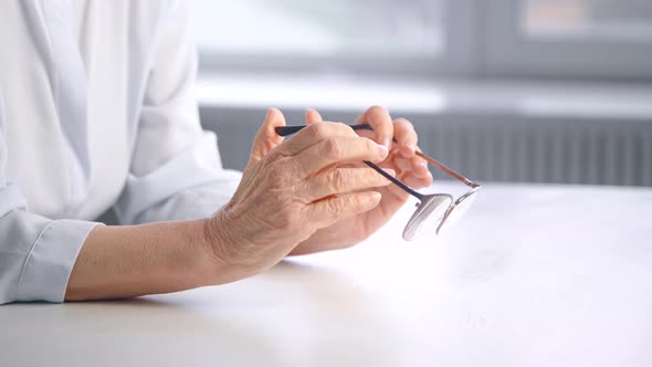 Grey haired old lady in white blouse holds glasses in hand with thoughtful wrinkly face