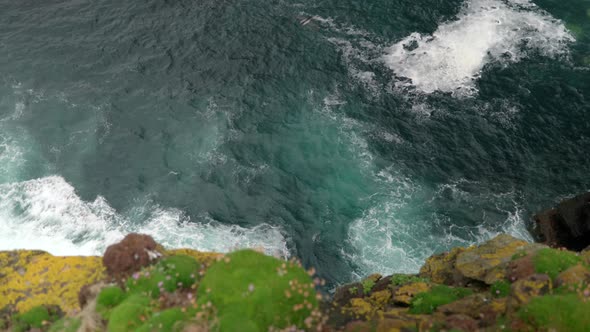 Waves gently crash over rocks and against the base of a sea cliff in the deep teal coloured ocean wh