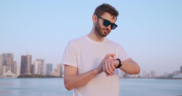 A Young Man Standing on the Waterfront in the Summer Uses the Smart Watch Screen