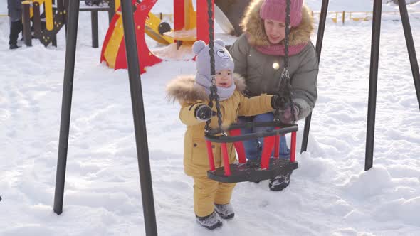 Mom shows curious newborn daughter a swing on a winter playground.