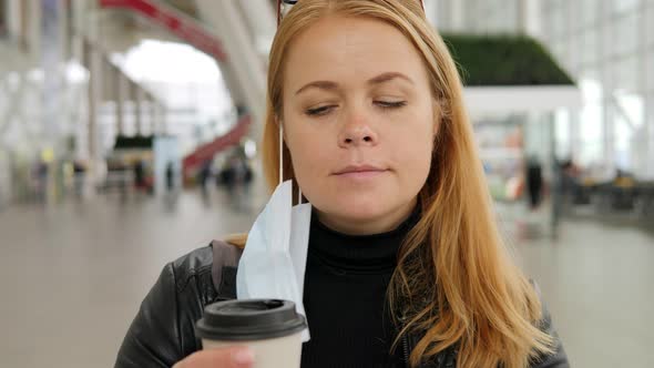 Portrait of a Young Woman in a Medical Mask at the Airport with a Cup of Coffee