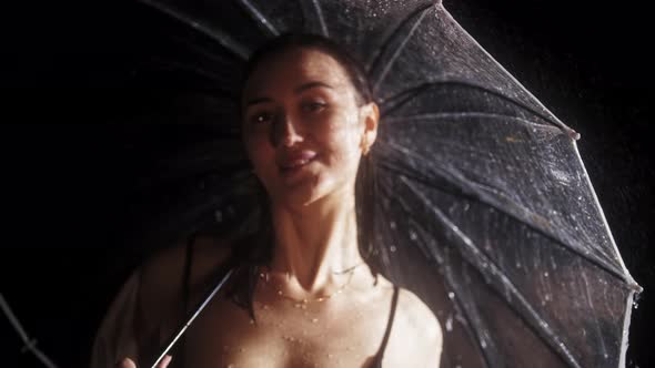 Young Flirty Woman in Black Swimming Suit Standing Under the Rain Holding an Umbrella and Looks at