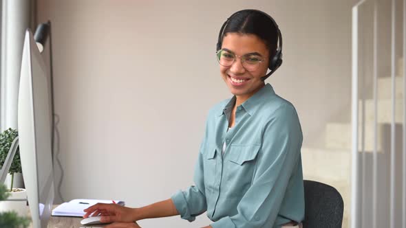 Young Cheerful Woman Using Headset for Online Talking Sitting at the Desk at Home Office