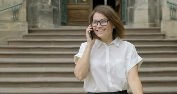 Mature Smiling Positive Woman in Glasses Light Blouse Walking on the Stairs Talking on the Phone