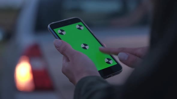 Woman Holding a Smartphone with Green Screen on Background of Broken Car