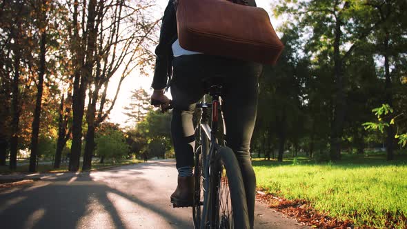 Young Businessman with Leather Bag Riding Bicycle to City Park Slow Motion Back View