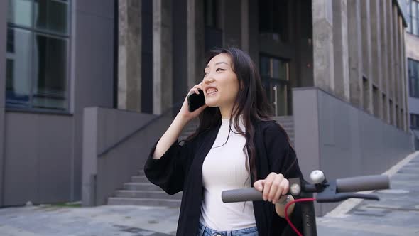 Asian Girl Going with Her E-Scooter Near Beautiful Building and Talking on Phone