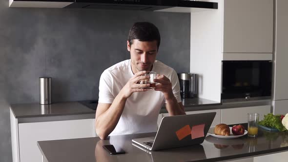 Portrait of a Handsome Relaxed Caucasian Man in a White Shirt Sitting on Kitchen in Front Him Laptop