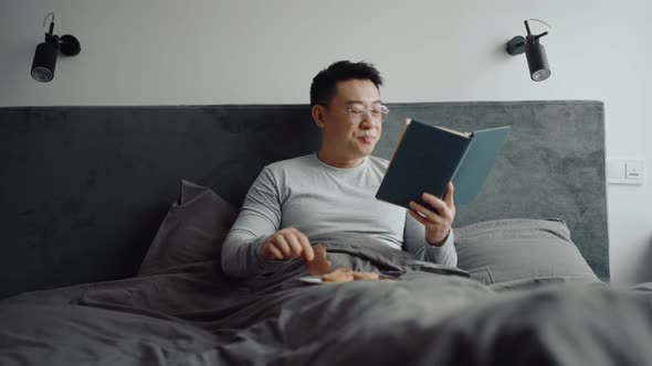 Happy Asian man wearing eyeglasses reading book and eating cookie