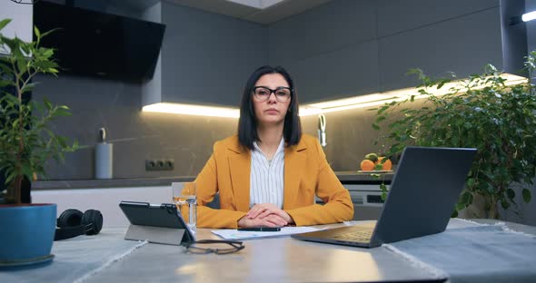 Woman in Glasses and Business Clothes Looking at Camera while Sitting at the Kitchen