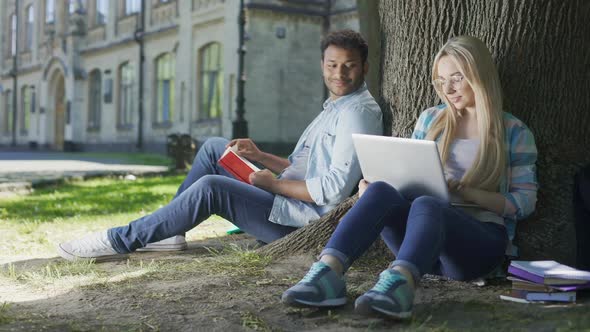 Young Man Sitting Under Tree With Book, Looking at Girl With Laptop, Indecisive