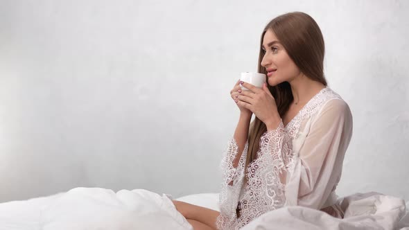 Smiling Morning Girl in Sexy Pajamas Drinking Coffee in Bed