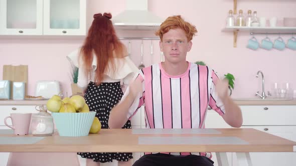 Portrait of Hungry Redhead Man Sitting at the Table in Kitchen Gesturing As Woman Cooking 