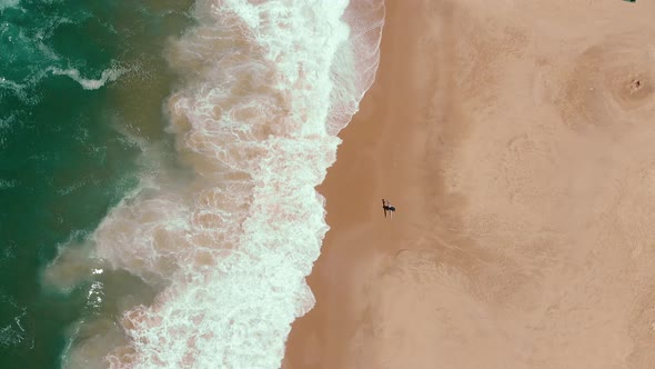 Top view of a lonely surfer walks on the beach while the wild endless sea crushing waves on the sand