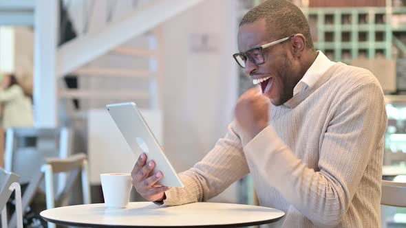 Excited African Man Celebrating Success on Tablet in Cafe