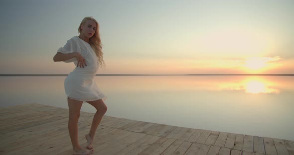 Pretty Model in White Dress is Dancing on the Pier During Colorful Sunset