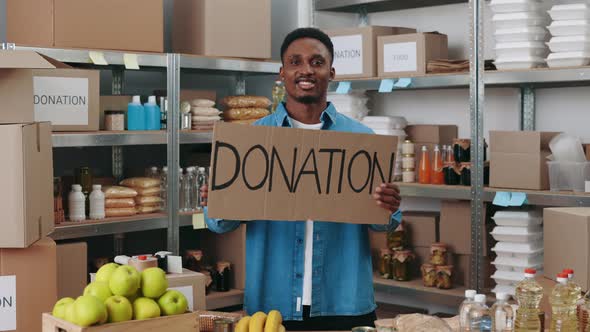 Smiling African American Male Volunteer in Casual Attire Holding Cardboard Banner with Work Donation
