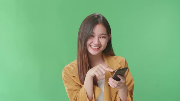 Smiling Woman Use Mobile Phone And Pointing To Smart Phone While Standing In The Green Screen Studio