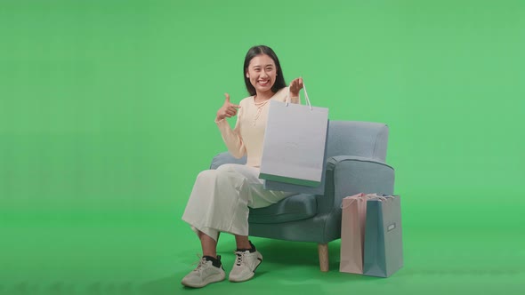 Woman Pointing Shopping Bags Before Smiling While Sitting On Sofa In Front Of Green Screen