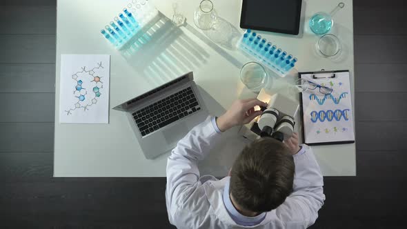 Lab Worker Looking Into Microscope, Recording Scientific Discovery on Laptop