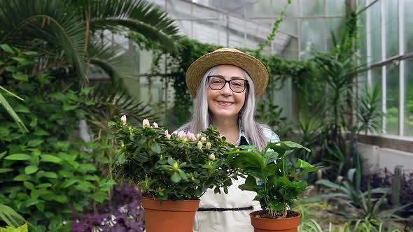 Senior Woman with Long Grey Hair in Hat which Holding Pots with Flowers and Posing on Camera