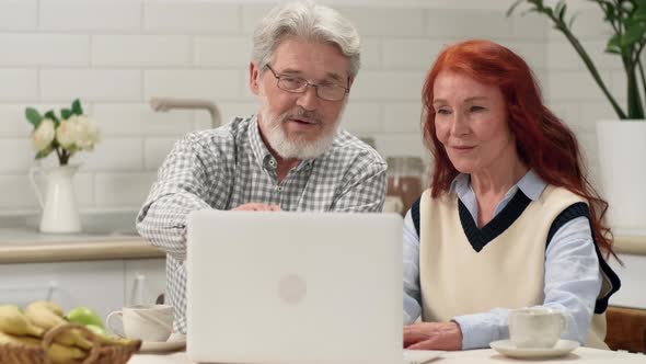 Happy Senior Couple in Their 60s and 70s Make a Video Call While Sitting at a Table in the Kitchen