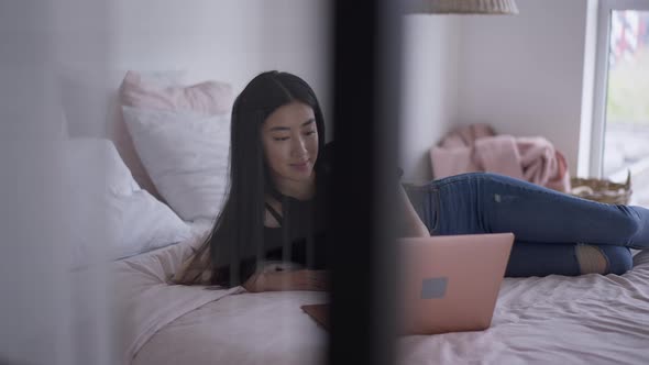 Charming Slim Asian Woman Lying on Bed Surfing Internet on Laptop